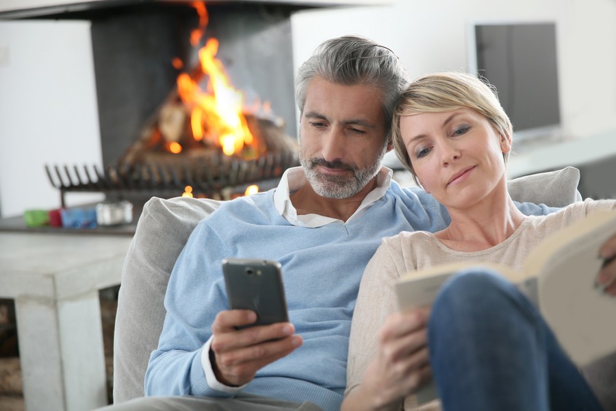 A middle-aged couple reclining on a couch looking at their phones with a stone fireplace in the background. 