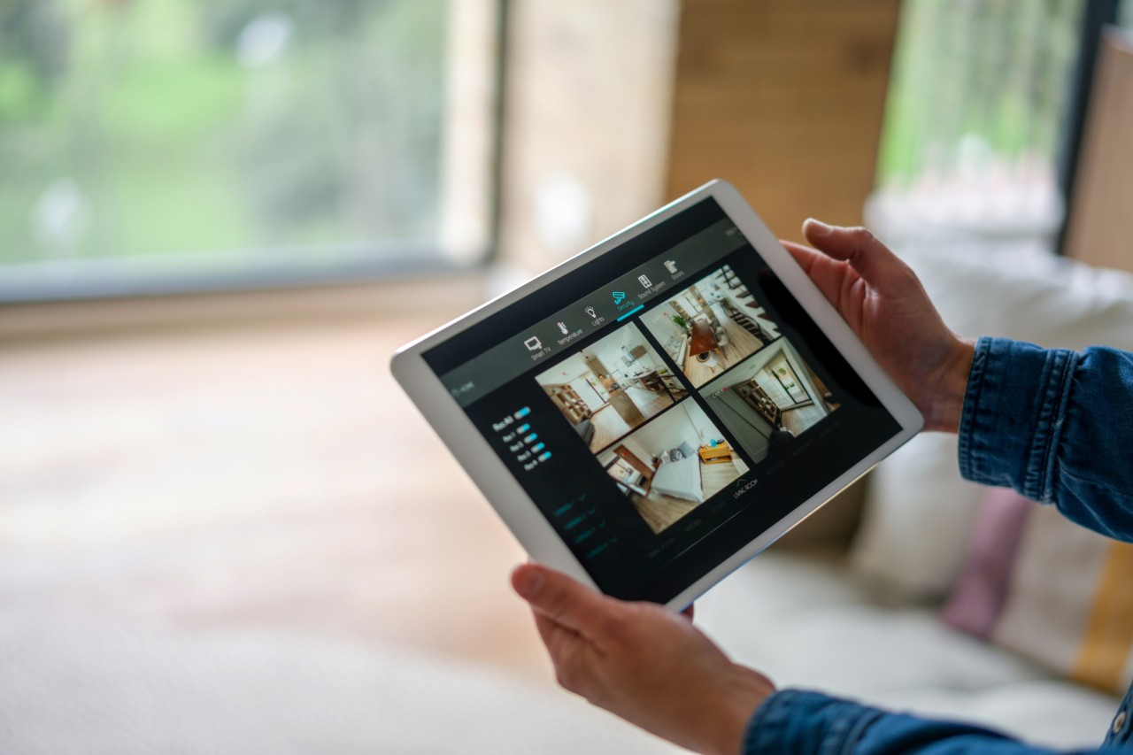A person holds a tablet displaying a home security interface with multiple live camera feeds, monitoring different rooms inside a modern home.